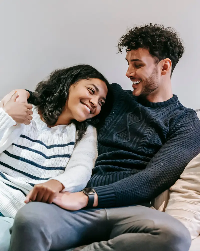 Smiling couple in knit sweaters relaxing together on a cream sofa in a bright room with light gray walls