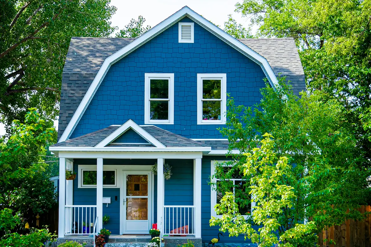 Blue gambrel-roof house with white trim and a white front porch, framed by tall trees