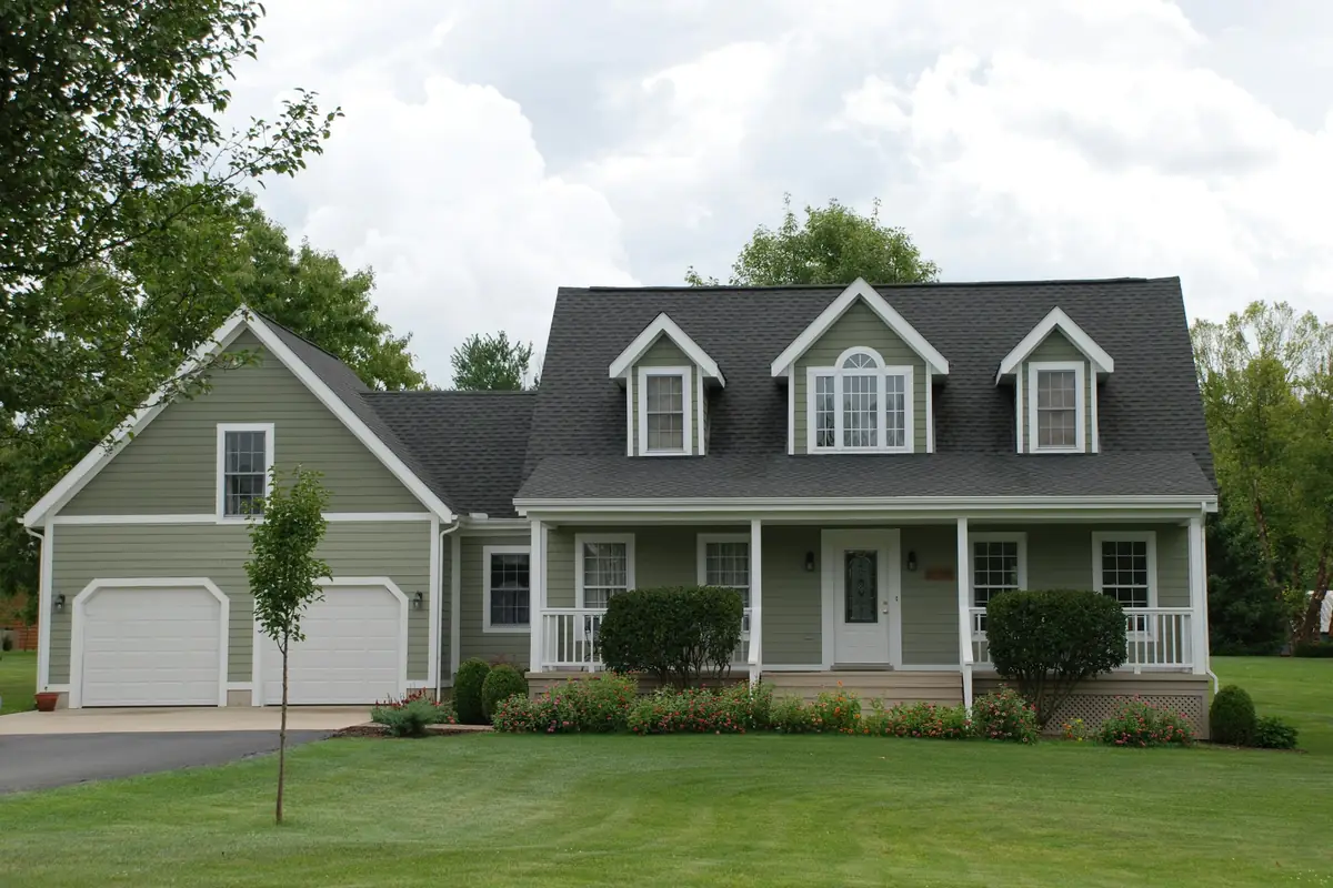 House with sage-green siding, white trim, three roof dormers, a covered front porch and a two-car garage above a wide lawn