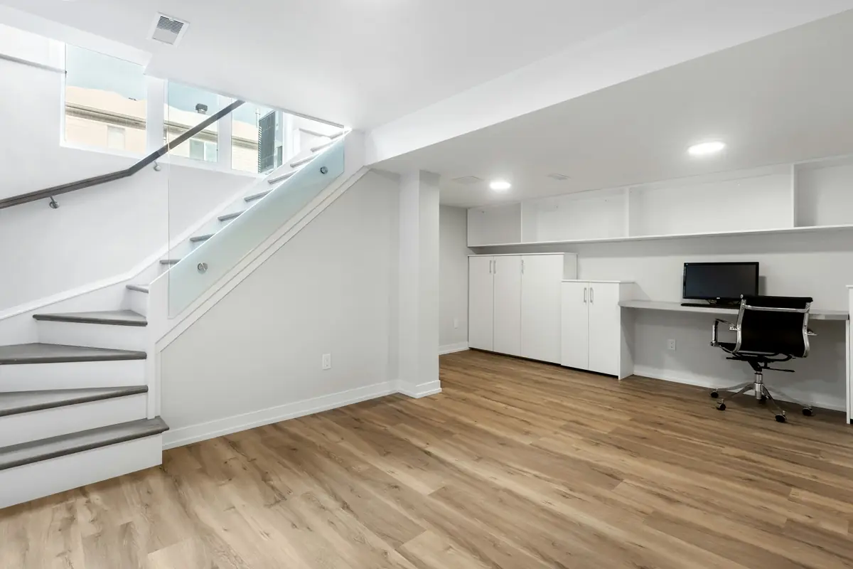 Finished lower level with white walls, a glass-paneled stair, white built-in cabinets and a desk on plank flooring