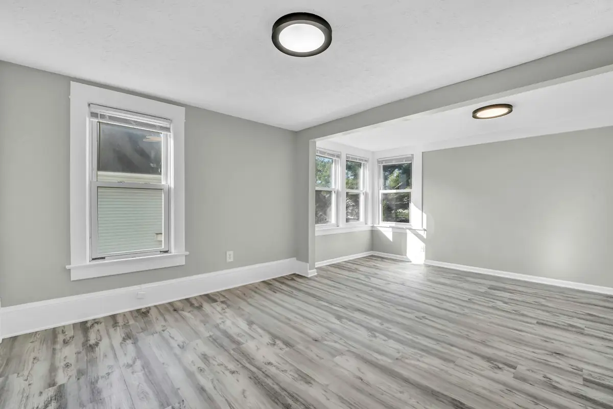 Empty room with light gray walls and gray plank flooring, opening onto a sunlit alcove with three windows