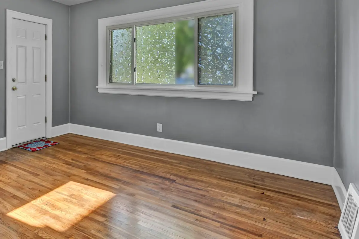 Empty room with mid-gray walls, white trim, a white six-panel door, a patterned-glass window and sunlight on a wood floor