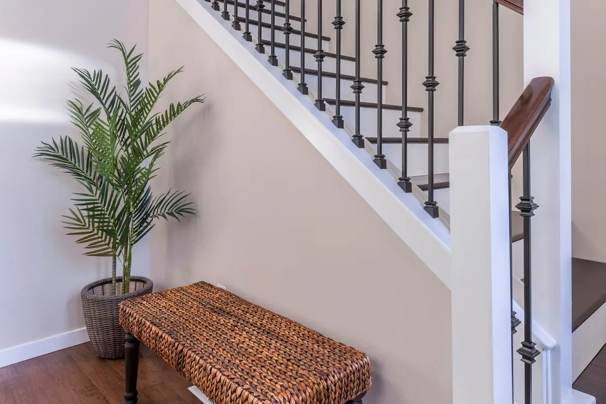 Staircase with white newel posts, black iron balusters and a wood handrail beside light taupe walls and a woven bench