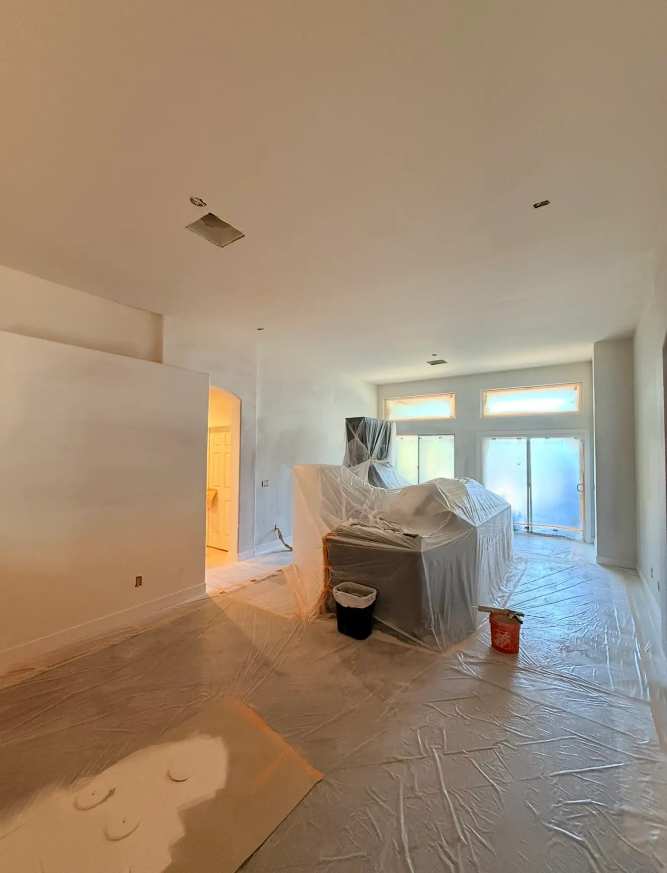 Large room masked for painting, with plastic over the floor, furniture, windows and sliding door, work in progress